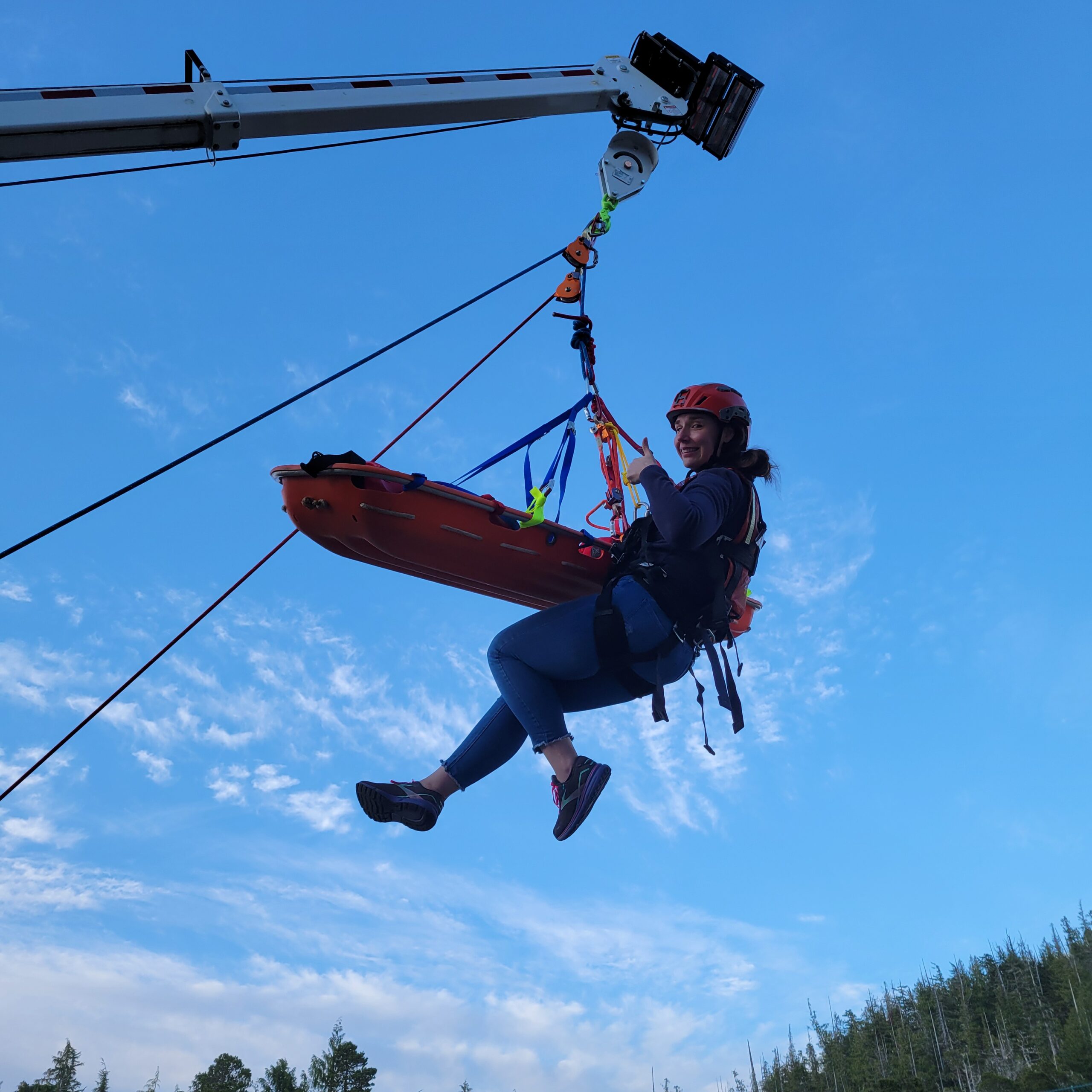 Person wearing a helmet and safety harness sits on a suspended zip-line chair against a bright blue sky, with forest below, mid-ride.